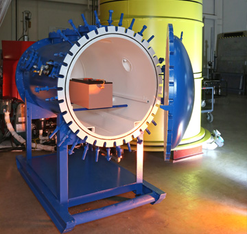 An altitude chamber within a battery test facility. It's a horizontal blue cylinder with an opening that opens towards us. 