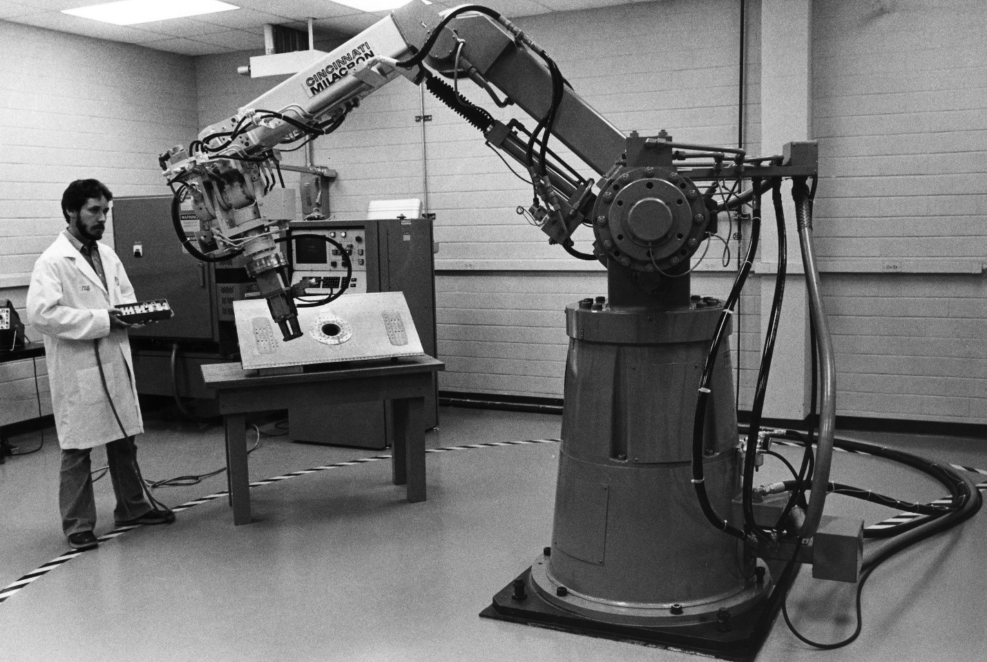 Black &amp; white photo showing a man in a lab coat standing near a large robotic arm