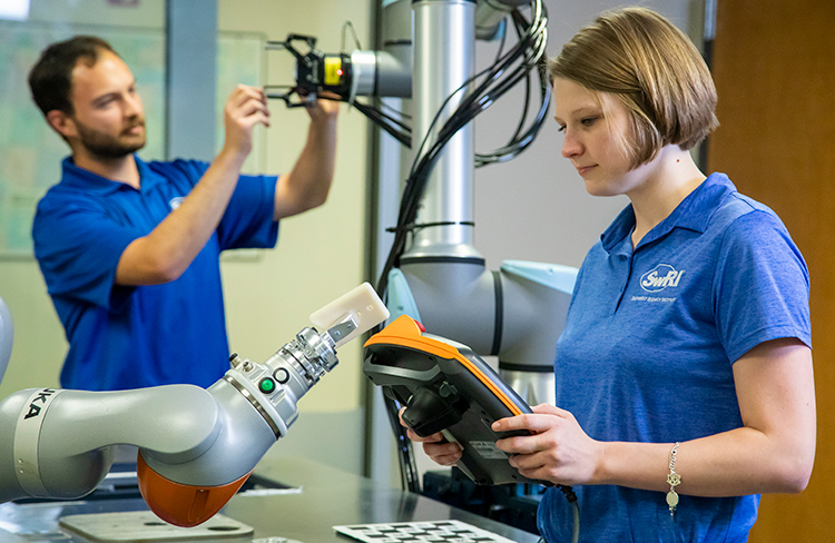 woman holding a cobot teach pendant in front of a cobot with a man in the background working on another robot.