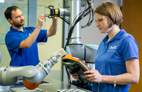 woman holding a cobot teach pendant in front of a cobot with a man in the background working on another robot