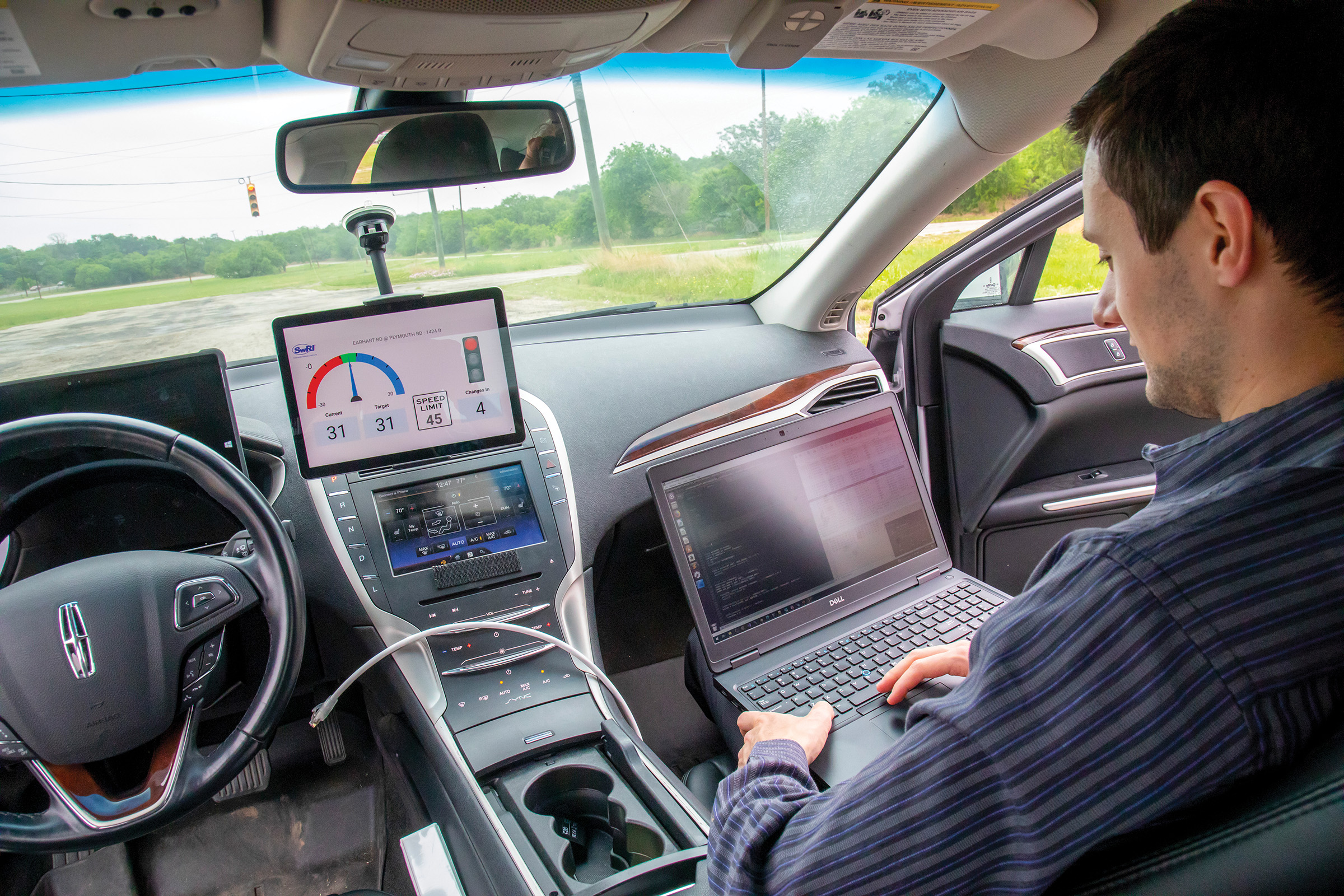 View from the backseat of NEXTCAR of a SwRI staff in passenger seat with laptop on lap and a view of the car's dashboard and windsheild
