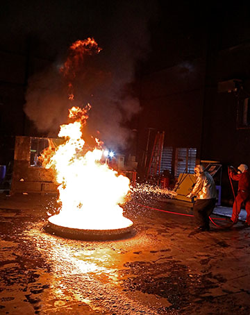 a large fire on the floor of a warehouse with a person spraying fire suppressing foam into it