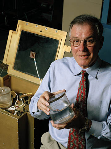 Dr. Franklin Dodge, in a blue shirt and red tie, holds a tank used in SwRI’s liquid motion experiment conducted onboard the space shuttle in 1997