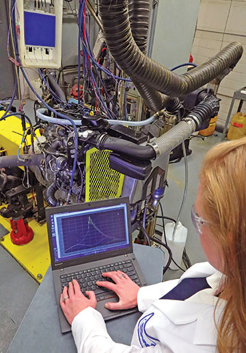 A lubricant testing lab with an engine on a test stand a female engineer reviewing data on a laptop