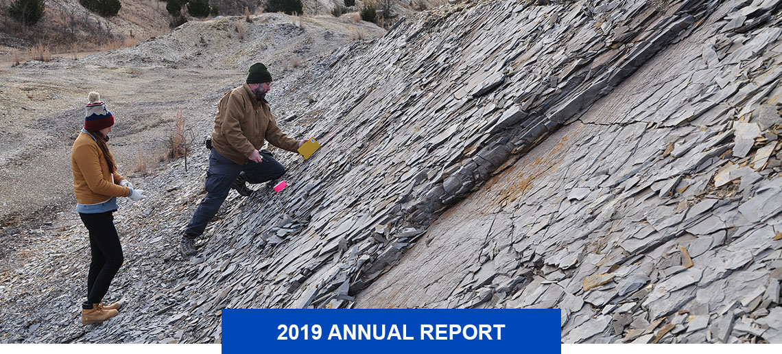 Outcrop with layers of rock flaking away and two people collecting specimens