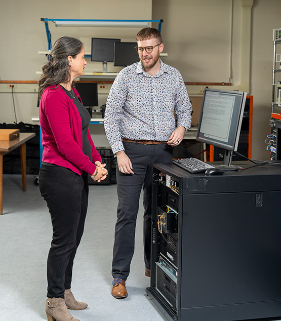 Two engineers standing near a computer system that's a part of the Programmable Military Waveform Environment Simulator.