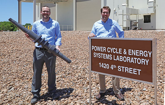 Aaron McClung holding a turbine shaft and Tim Allison standing behind a brown building sign