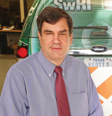 White man in dress shirt and tie sitting in front and to the left of the rear-end of a SUV