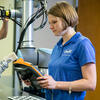 woman holding a cobot teach pendant in front of a cobot with a man in the background working on another robot.