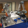 an orange and silver cobot sitting on work bench to the left of a blue and silver cobot picking up widget in gripper