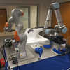 an orange and silver cobot sitting on work bench to the left of a blue and silver cobot picking up widget in gripper