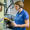 woman holding a cobot teach pendant in front of a cobot with a man in the background working on another robot