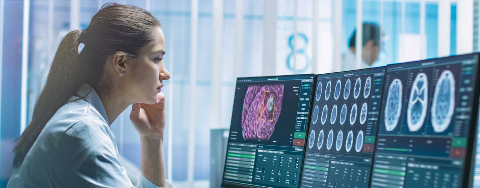 woman in lab coat looking at computer monitors displaying various brain scans