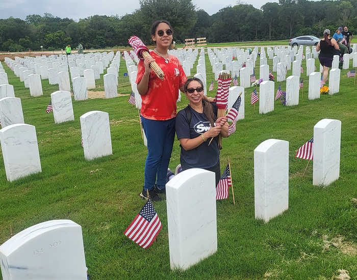 two hispanic women, one standing, one kneeling, behind white granite headstones, holding american flags