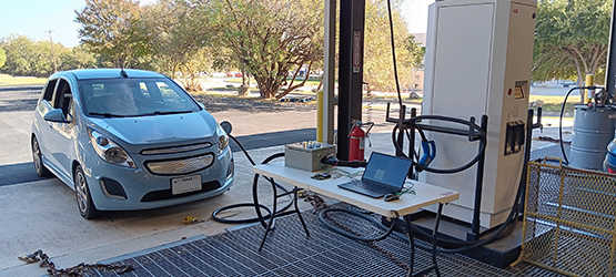 Blue electric car being charged, next to table with laptop. SwRI performs cybersecurity analysis of direct-current, fast-charging for electric vehicles. 