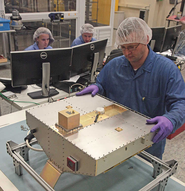 man in protective equipment building microsatellite while 2 people in the background work on computers