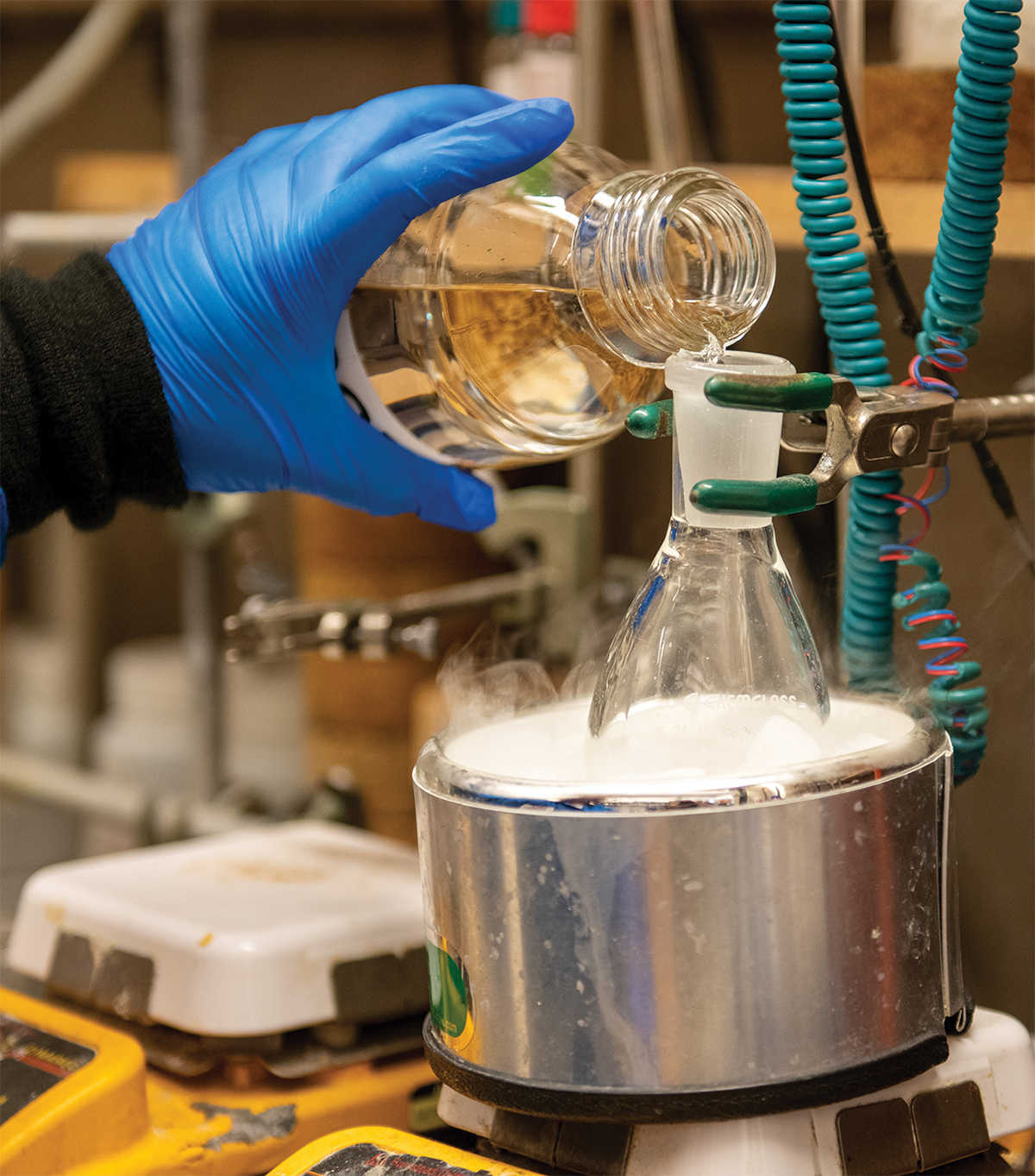 blue-gloved hand pouring chemical into heated beaker