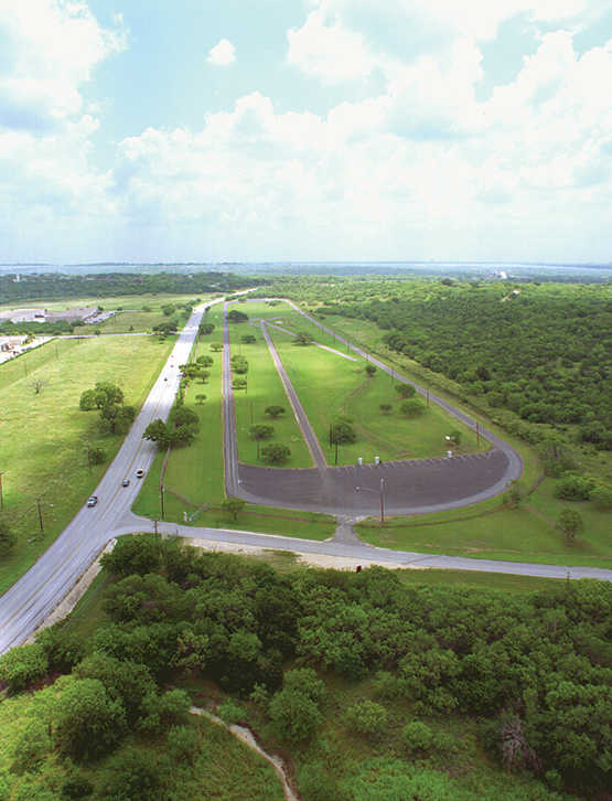 High up drone view of a test track road, surrounded by green grass and trees.