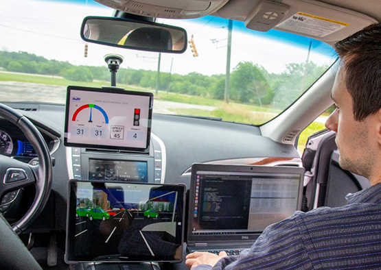 Man sitting in passenger seat inside a car, staring at the dashboard where dual ipad screens show SwRI software. He also has a laptop on his lap. 
