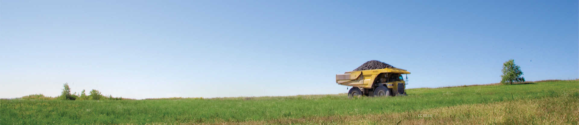A landscape with green grass and a blue sky day with a yellow hauling truck carrying rocks cruising through the field.