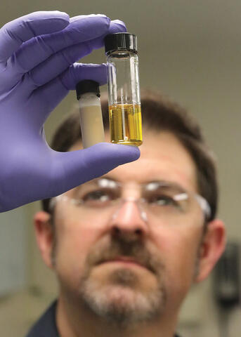 in the foreground a man's purple gloved hand holding 2 test tubes between his thumb and ring/index fingers. in background a man wear safety glasses staring at the test tubes
