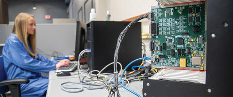 white woman with blonde hair in blue clean suit, working at a computer in the background. in foreground is a microprocessor connected to computer