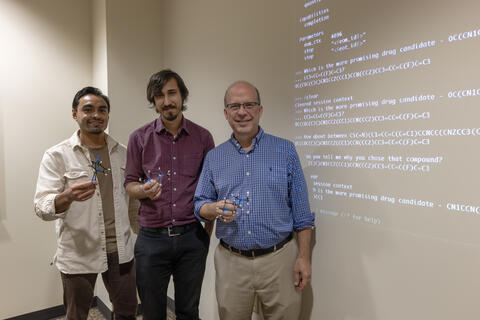 Photograph of Research Scientist Daniel Hinojosa, Lead Computer Scientist Michael Hartnett and Staff Scientist Dr. Johnathan Bohmann holding up a visual representation of a common molecule used for the synthesis of pharmaceuticals