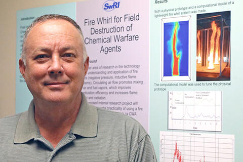 white man in gray polo shirt standing in front of a poster than reads Fire Whirl for Field Destruction of Chemical Warfare Agents