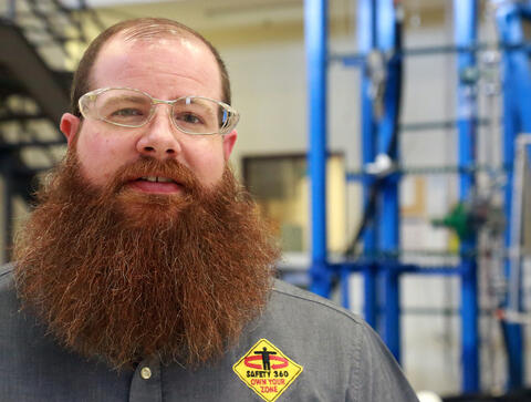 Matthew Herron, with a beard and safety glasses, standing in front of scaffolding