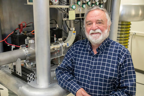 older white man with beard wearing blue shirt sitting in front of space instrument