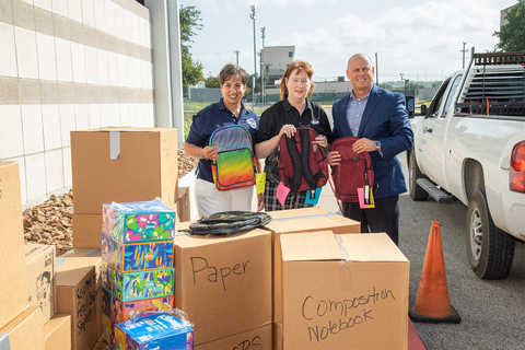 two women and 1 man holding backpacks standing behind cardboard boxes labeled with types of school supplies 