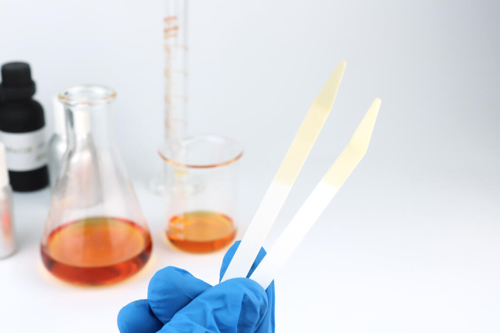 Blotter papers in a right hand with background essential oil and fragrance oil bottle for testing smell during blending process for choosing nice scent for scented candles and body perfume in lab