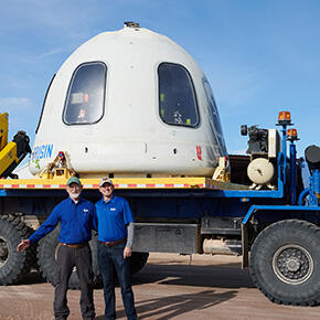 Engineer Kevin Supak and engineer Steven Green standing in front of part of Blue Origin’s New Shepard vehicle