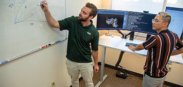 one chemist drawing on white board while second chemist in front of computer looks on