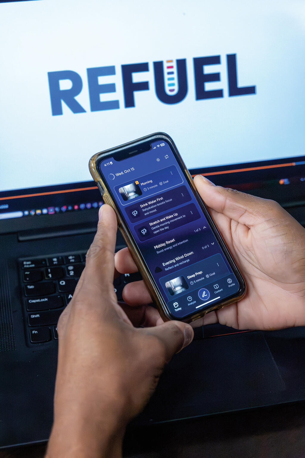 Image of laptop with REFUEL logo on screen with person holding a cell phone in the foreground with REFUEL mobile app open