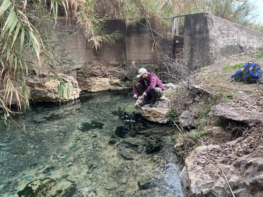 SwRI Research Scientist Dr. Dylan Parmenter leaning over a spring feeding the Rio Grande River to  monitor the water