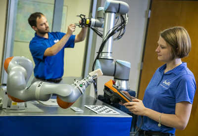 In foreground, a woman using a control pad to manipulate a collaborative robot; in background, a man is working on a second robot